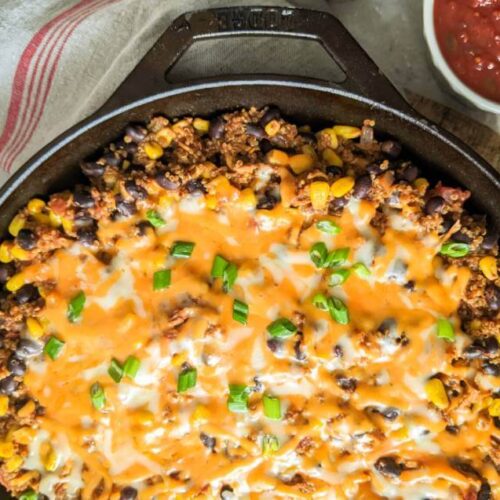 A cast iron skillet filled with baked casserole topped with melted cheese, black beans, corn, and chopped green onions, with a bowl of salsa on the side.