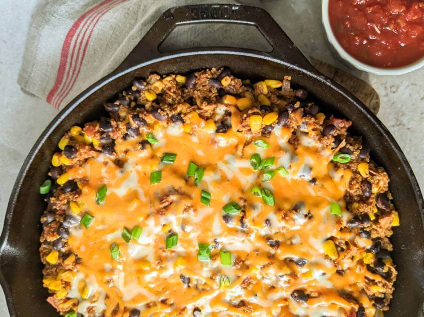 A cast iron skillet filled with baked casserole topped with melted cheese, black beans, corn, and chopped green onions, with a bowl of salsa on the side.
