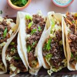 Five soft tacos filled with Korean beef, shredded lettuce, green onions, and sesame seeds are arranged on a tray. Small bowls with herbs and sesame seeds are nearby.