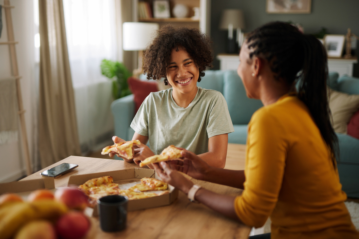teenager is a picky eater eating with mom with a box of pizza on the table and smiling at each other