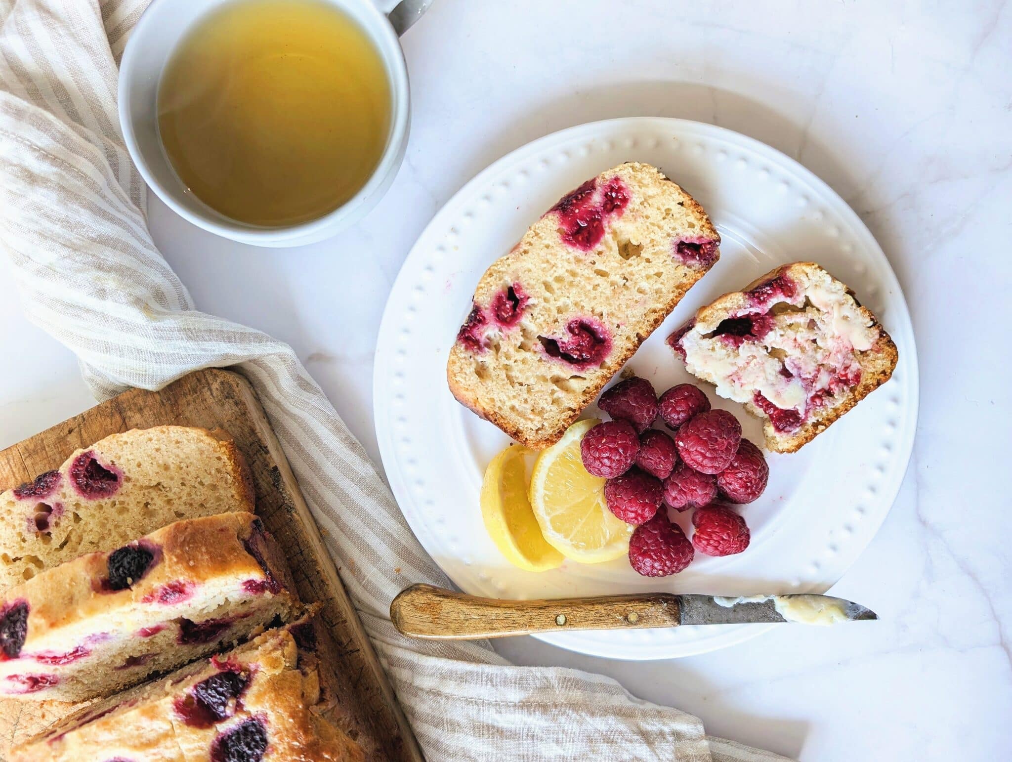 top down raspberry lemon loaf in slices with butter on a white plate topped with raspberries and lemon slices