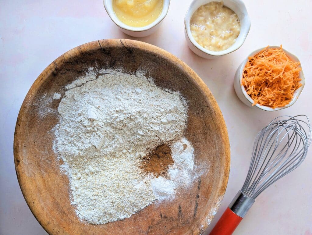 flour in a mixing bowl with wet ingredients measured out on the side including carrots and banana